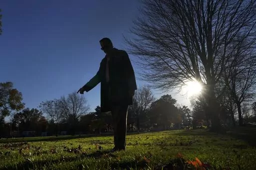 Cook County, Ill., Sheriff's Commander Jason Moran, who leads the sheriff's missing persons initiative stands in silhouette and points to a round cylinder that marks a grave of a person who self-identified as Seven, at the Mount Olivet Cemetery on Chicago's Far South Side Monday, Nov. 13, 2023. “That’s a horrible circumstance that someone could die and no one knows who they are. That’s why we pursue these cases so strongly, out of dignity,” says Moran, who oversees the sheriff’s missin