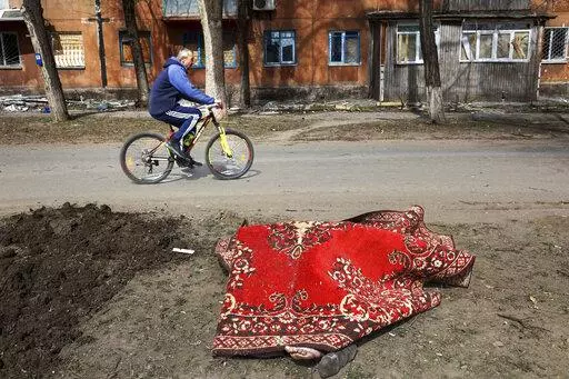 A man rides his bicycle past a body covered by a rug after fighting on the outskirts of Mariupol, Ukraine, in territory under control of the separatist government of the Donetsk People's Republic, on Tuesday, March 29, 2022. (AP Photo/Alexei Alexandrov)