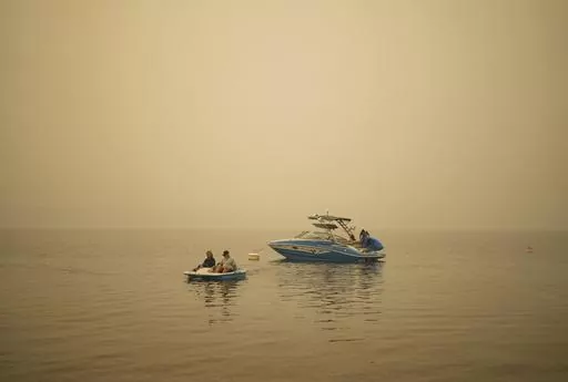 Smoke fills the air as Pat Manzuik and her husband Trevor use a paddleboat to get to shore after being given a boat ride by good samaritan Christy Dewalt, back right, back to their home they were evacuated from due to the Lower East Adams Lake wildfire, in Scotch Creek, Canada, Sunday, Aug. 20, 2023. (Darryl Dyck/The Canadian Press via AP)