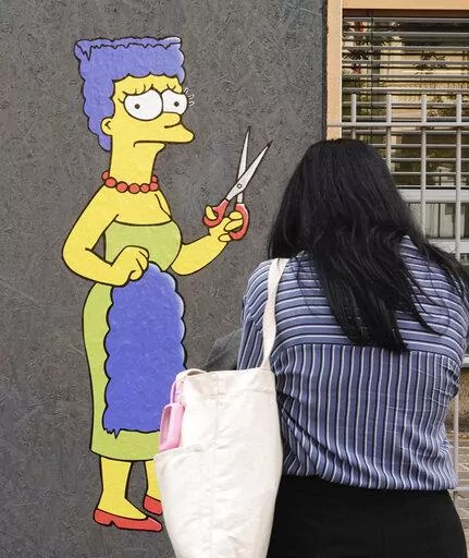 A woman takes pictures of a mural called "The Cut" by street artist aleXsandro Palombo depicting Marge Simpson, a character of the animated television series "The Simpsons" cutting her iconic hair, in front of the Consulate of Iran, in Milan, Italy, Wednesday, Oct. 5, 2022. Thousands of Iranians have taken to the streets over the last two weeks to protest the death of Mahsa Amini, a 22-year-old woman who had been detained by Iran's morality police in the capital of Tehran for allegedly not adher